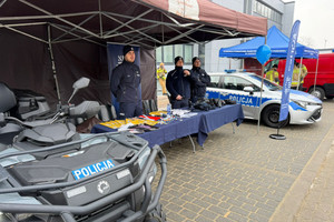 Na zdjęciu widoczni trzej umundurowani policjanci przy stoisku profilaktycznym. Widoczny też radiowóz i policyjny quad.