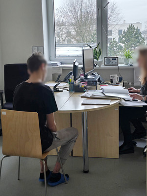 Na zdjęciu widoczny mężczyzna, który siedzi na krześle. Przy biurku nieumundurowana policjantka.