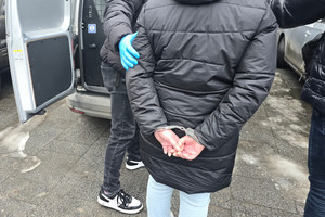 Na zdjęciu widoczna kobieta z założonymi na ręce trzymane z tyłu kajdankami, która jest prowadzona przez nieumundurowanego policjanta.