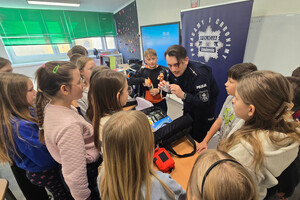 Na zdjęciu widoczny umundurowany policjant, który przedstawia sprzęt ratunkowy uczniom, stojącym wokół niego.