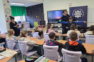 Na zdjęciu widoczny umundurowany policjant, który pomaga ściągnąć uczniowi policyjną kamizelkę. Widoczni także uczniowie siedzący w ławkach.