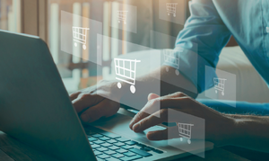 Zdjęcie przedstawia częściowo widocznego mężczyznę, który siedzi przy laptopie. Widoczne są także grafiki w postaci wózków sklepowych. Zdjęcie z portalu cert.pl