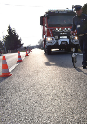 Na zdjęciu widoczny policjant ruchu drogowego, wóz strażacki oraz pachołki rozstawione na jezdni.