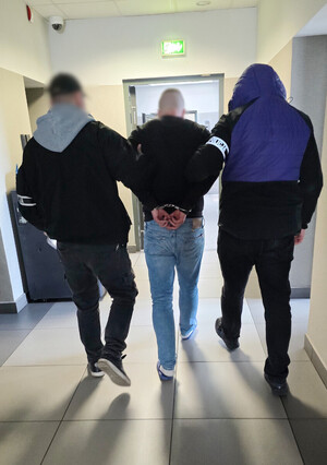 Dwaj nieumundurowani policjanci prowadzą zatrzymanego mężczyznę. Mężczyzna ma założone kajdanki na ręce trzymane z tyłu.