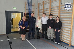 Policjant, dwie kobiety oraz dwóch uczniów i uczennica stoją na sali gimnastycznej.