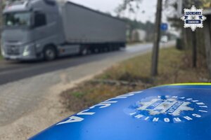Widoczna maska radiowozu oraz jadący drogą samochód ciężarowy.