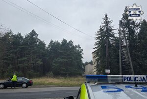 Częściowo widoczny radiowóz, a po drugiej stronie ulicy policjant w trakcie kontroli drogowej pojazdu.
