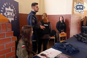 Policjant mówi o kamizelce policyjnej, którą ma ubrana dziewczyna stojąca przed nim. Widoczna są też dwie dziewczyny siedzące przy stoisku profilaktycznym.