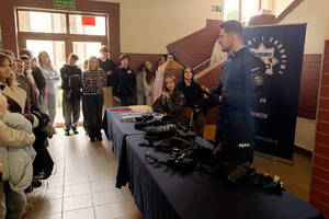 Policjant przy stoisku profilaktycznym podczas Dnia Otwartego szkoły. Wokół zgromadzeni są uczniowie. Na stolikach leżą materiały profilaktyczne oraz sprzęt policyjny.