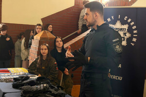 Policjant przy stoisku profilaktycznym podczas Dnia Otwartego szkoły. Wokół zgromadzeni są uczniowie. Na stolikach leżą materiały profilaktyczne oraz sprzęt policyjny.