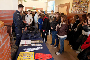 Policjant przy stoisku profilaktycznym podczas Dnia Otwartego szkoły. Wokół zgromadzeni są uczniowie. Na stolikach leżą materiały profilaktyczne oraz sprzęt policyjny.