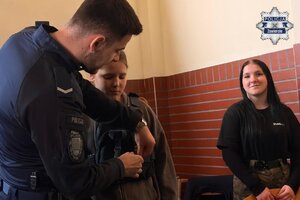 Policjant mówi pomaga zapiąć dziewczynie kamizelkę policyjną. Obok stoi nastolatka.