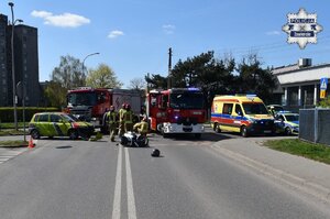 Miejsce wypadku drogowego. Na jezdni stoi samochód osobowy z roztrzaskanym przodem, a po jego prawej stronie leży motocykl. W tle widoczne pojazdy służbowe straży pożarnej, karetkę pogotowia oraz radiowóz, a także strażacy wykonujący czynności.