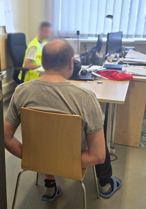Zatrzymany mężczyzna siedzi na krześle w trakcie przesłuchania. Przy biurku widoczny nieumundurowany policjant w kamizelce odblaskowej.