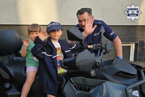 Dwójka dzieci na quadzie i stojący obok policjant machają dłońmi.