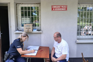 Przy stoliku przed budynkiem siedzi policjantka i sporządza dokumentację. Na przeciwko niej siedzi mężczyzna.