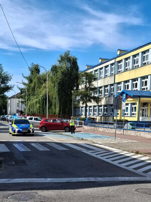 Policjantka przeprowadza lustrację terenu przed szkołą. Widoczny także radiowóz.